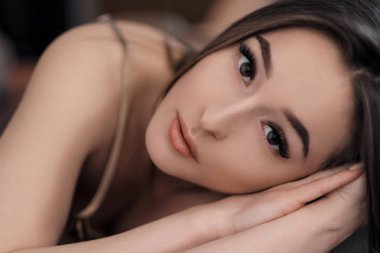 Çıplak omuzları yatakta yatan kahverengi gözlü güzel esmer kız. Yakından yüzleşin. Asya ve Doğu tipi görünüm.