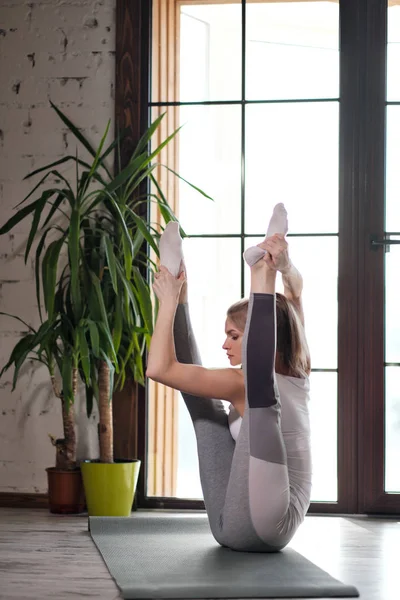 Jovem bela mulher atlética loira em sportswear branco fazendo ioga e alongamento em um tapete cinza no fundo de uma grande janela de edifício de vários andares . — Fotografia de Stock