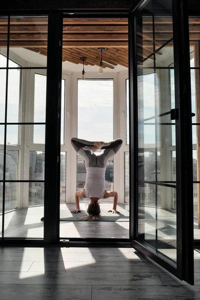Jovem bela mulher atlética loira em sportswear branco fazendo ioga e alongamento na varanda em casa. Executa headstand . — Fotografia de Stock