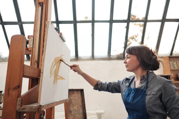 Joven mujer hermosa pintor entre caballetes y lienzos en un estudio brillante . — Foto de Stock