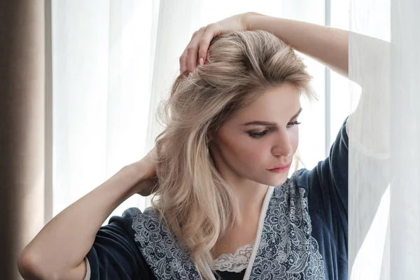 Mooie jonge vrouw blonde in een blauwe gewaad door het raam. — Stockfoto