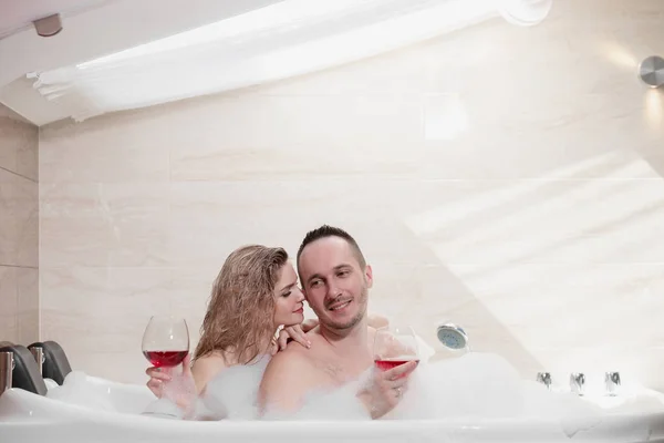 An attractive couple, a man and a woman, a husband and wife, lie in a bubble bath and drink red wine. — Stock Photo, Image