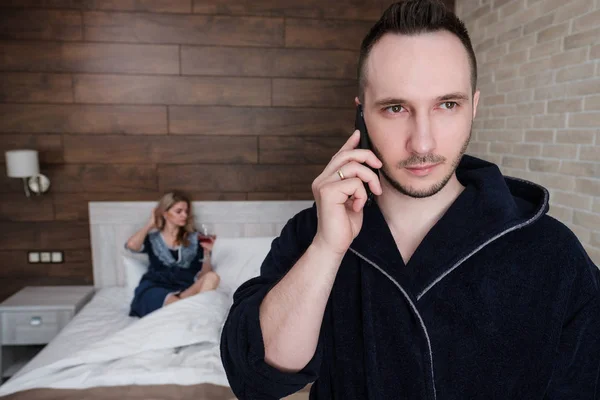 Couple, man and woman in the hotel room, in the bedroom. The husband stands at the window and speaks on the phone, the wife lies on the bed and waits for him.