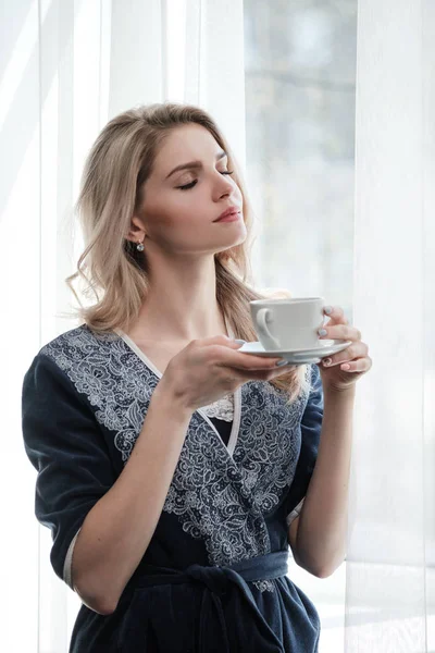 Krásná mladá blondýnka v modrém rouchu u okna. Káva nebo čaj se pije z bílého šálku s talířem. Dobré ráno, sluníčko, okno ložnice. — Stock fotografie