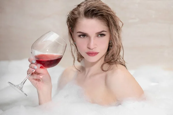 Atrakcyjna blond kobieta leżąca w wannie w piance w hotelu. Picie czerwonego wina z dużego szklany kieliszek. — Zdjęcie stockowe