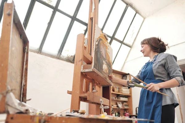 Mulher artista pinta um quadro em tela. Estúdio de arte brilhante com uma grande janela. Bagaços e telas . — Fotografia de Stock