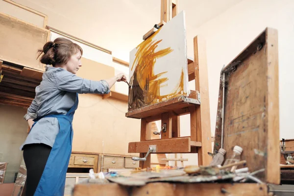 Joven mujer hermosa pintor entre caballetes y lienzos en un estudio brillante . — Foto de Stock