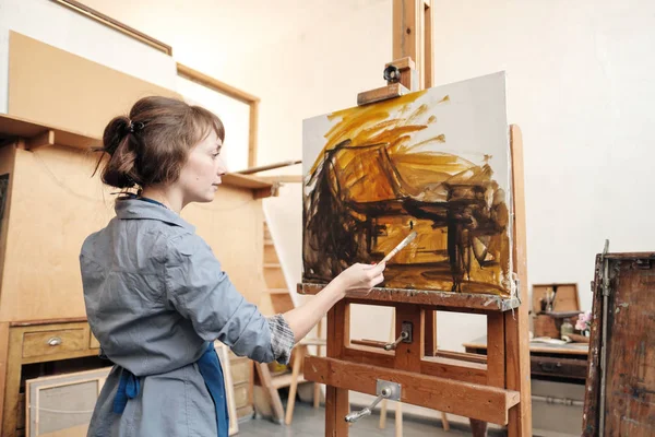 Joven mujer hermosa pintor entre caballetes y lienzos en un estudio brillante . — Foto de Stock