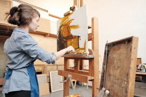 Joven mujer hermosa pintor entre caballetes y lienzos en un estudio brillante . — Foto de Stock