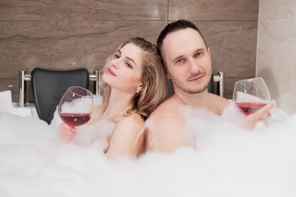 Um casal atraente, um homem e uma mulher, um marido e uma esposa, deitam-se num banho de espuma e bebem vinho tinto. . — Fotografia de Stock