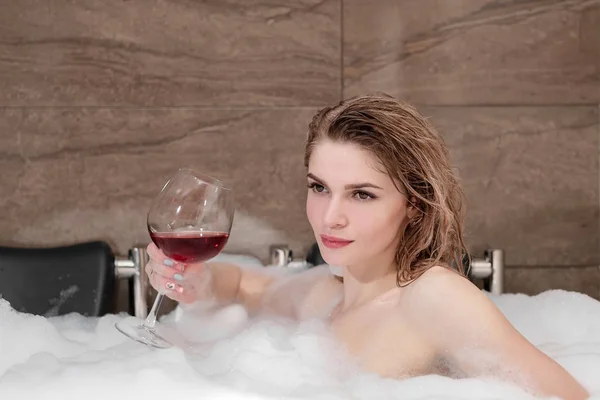 迷人的金发女郎躺在浴缸泡沫在酒店。从大玻璃杯中喝红酒. — 图库照片