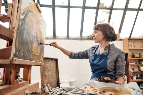 Woman artist paints a picture on canvas. Bright art studio with a large window. Easels and canvases. — Stock Photo, Image