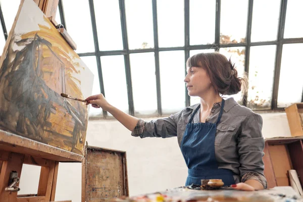 Mulher artista pinta um quadro em tela. Estúdio de arte brilhante com uma grande janela. Bagaços e telas . — Fotografia de Stock