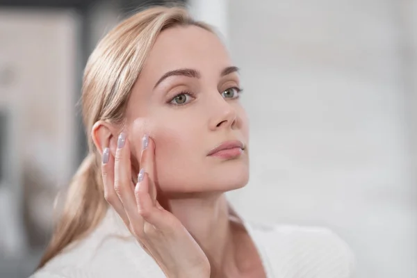 Beyaz bornozlu orta yaşlı sarışın kadın aynanın yanında banyoda duruyor. Cilde dokunuyor ve gülümsüyor..