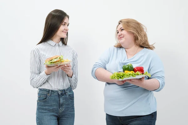 Deux filles, épaisses et minces, avec des aliments sains à base de légumes et de fruits et un fast-food malsain avec un hamburger. Le concept de régime alimentaire . — Photo