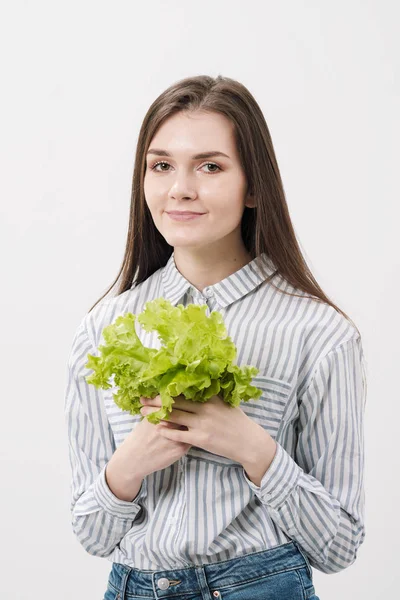 Une fille brune mince avec les cheveux longs sur un fond blanc, tient dans ses mains et montre les feuilles de laitue fraîche verte . — Photo