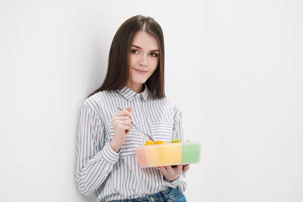 Chica morena delgada con el pelo largo sobre un fondo blanco come con un tenedor de un recipiente para almuerzos. Caja de color para alimentos, una selección de diferentes productos y un almuerzo completo . —  Fotos de Stock