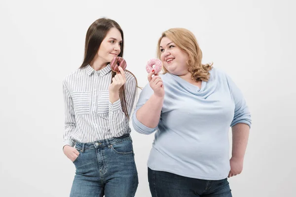 Two girls, thin and fat on a white background, are holding pink glazed donuts in their hands.