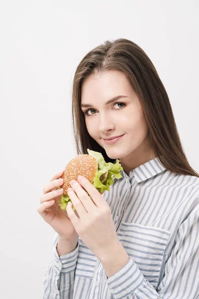 Young beautiful brunette girl on a white background with a hamburger in her hands. — Stock Photo, Image