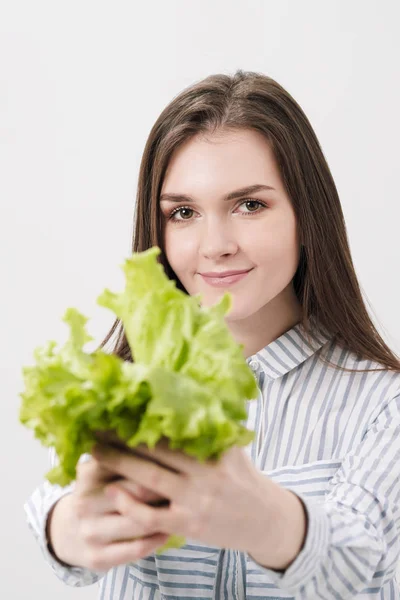 A slender brunette girl with long hair on a white background, holds in her hands and shows the leaves of green fresh lettuce. — Stock Photo, Image