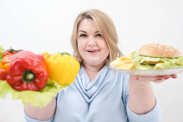 Jeune femme de graisse luxuriante dans des vêtements bleus décontractés sur un fond blanc tenant une salade de légumes et une assiette de restauration rapide, hamburger et frites. Alimentation et bonne nutrition . — Photo