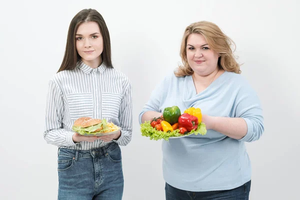 Duas meninas, grossas e magras, com alimentos saudáveis de legumes e frutas e fast food insalubre com um hambúrguer. O conceito de dieta . — Fotografia de Stock