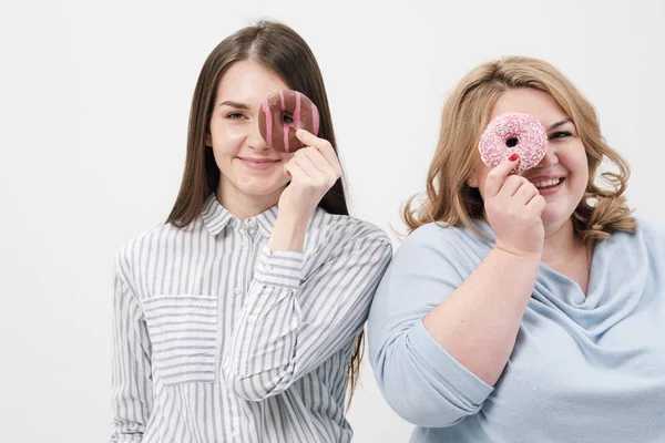 Two girls, thin and fat on a white background, are holding pink glazed donuts in their hands.