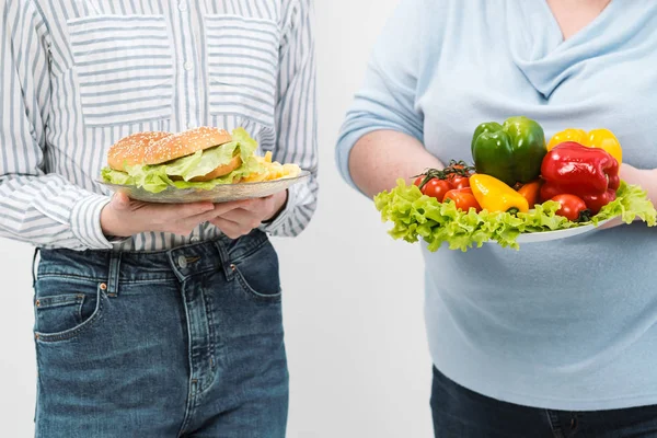 Dos chicas, gruesas y delgadas, con comida saludable de verduras y frutas y comida rápida poco saludable con una hamburguesa. El concepto de dieta . — Foto de Stock
