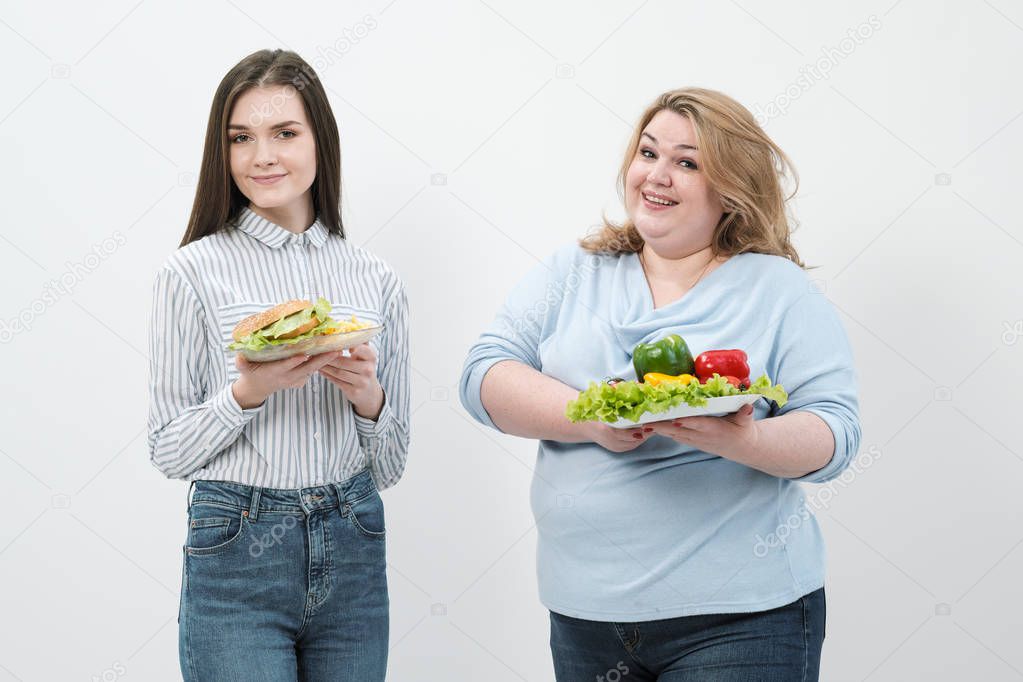 Two girls, thick and thin, with healthy food from vegetables and fruits and unhealthy fast food with a hamburger. The concept of diet.