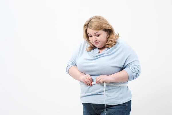 Şişman bir kadın beyaz bir arka plan üzerinde rahat giyim bir ölçüm bandı ile bel ölçer.