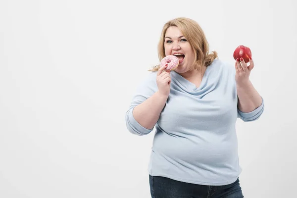 Uma mulher loira gorda e gorda em um fundo branco, segurando donuts de vidro rosa e uma maçã vermelha . — Fotografia de Stock