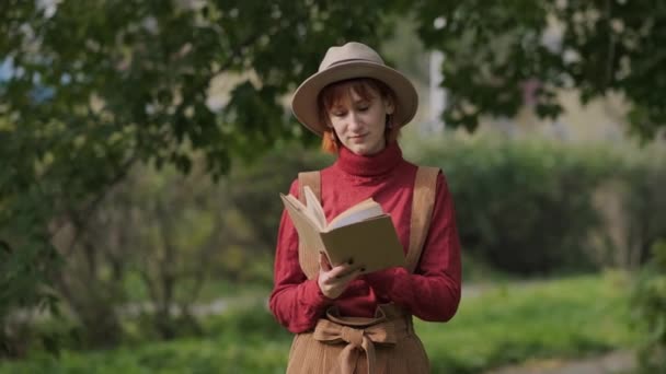 Jonge aantrekkelijke roodharige vrouw met hoed en trui leesboek in een natuurpark. Herfst koele dag. — Stockvideo
