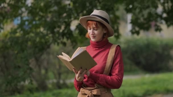 Joven mujer pelirroja atractiva en sombrero y suéter libro de lectura en un parque natural. Día fresco de otoño . — Vídeo de stock