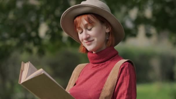 Jonge aantrekkelijke roodharige vrouw met hoed en trui leesboek in een natuurpark. Herfst koele dag. — Stockvideo