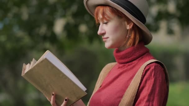 Jonge aantrekkelijke roodharige vrouw met hoed en trui leesboek in een natuurpark. Herfst koele dag. — Stockvideo