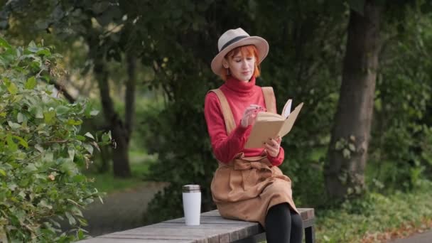 Junge attraktive rothaarige Frau mit Hut und Pullover, die auf einer Bank im Naturpark ein Buch liest und Kaffee aus einer Thermobecher trinkt. Herbstlicher kühler Tag. — Stockvideo