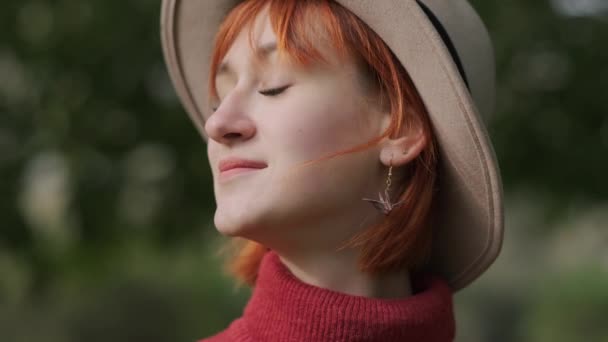 Młoda atrakcyjna rudowłosa dziewczyna z krótkimi włosami w kapeluszu i swetrem z terakoty w parku naturalnym w jesienny dzień. — Wideo stockowe
