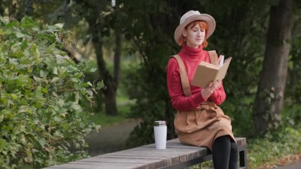 Mulher ruiva atraente nova em um chapéu e suéter que lê um livro e bebe o café de um thermocup em um banco em um parque natural. Outono dia fresco . — Vídeo de Stock