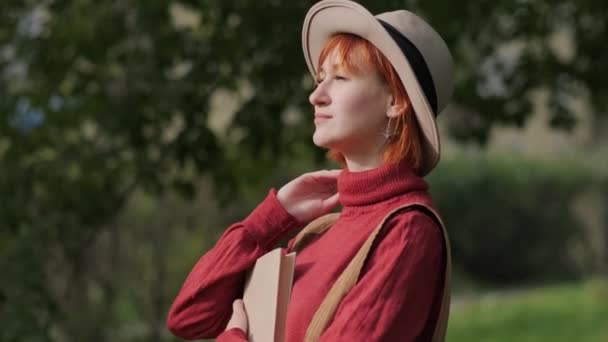 Giovane donna rossa attraente con cappello e maglione che legge un libro su una panchina in un parco naturale. Autunno giornata fresca . — Video Stock