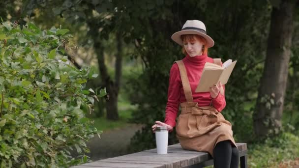 Giovane donna rossa attraente con cappello e maglione che legge un libro e beve caffè da una termocoppa su una panchina in un parco naturale. Autunno giornata fresca . — Video Stock