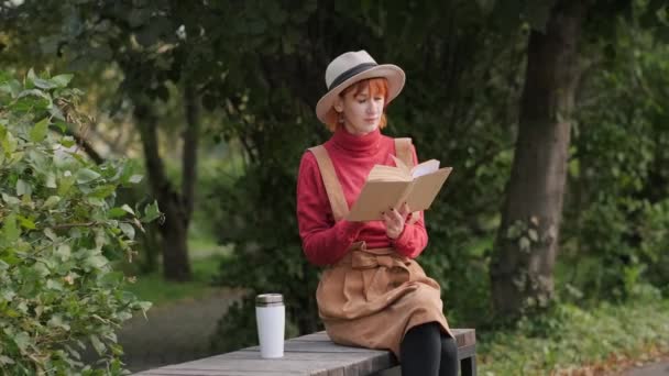 Молодая привлекательная рыжая женщина в шляпе и свитере читает книгу и пьет кофе из термокапа на скамейке в природном парке. Осенний прохладный день . — стоковое видео