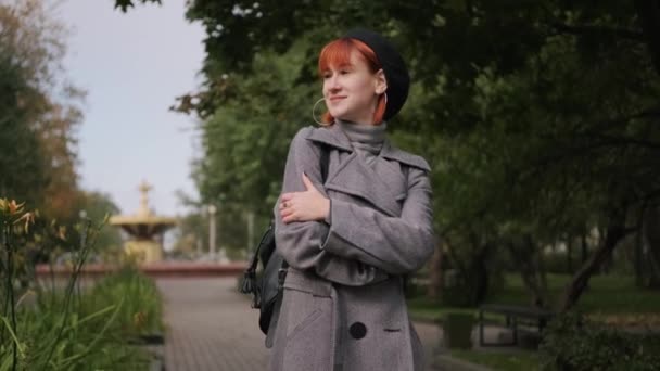 Jong aantrekkelijk roodharig meisje met kort haar in een zwarte baret en grijze jas in een natuurpark op een herfstdag. — Stockvideo
