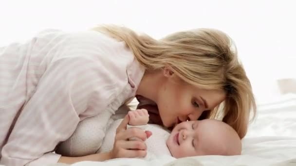 Mladá matka blondýna v pyžamu objetí a stará se o malou holčičku v jezdci. Matka a dítě leží na posteli s bílým povlečením u velkého okna se závěsem. — Stock video