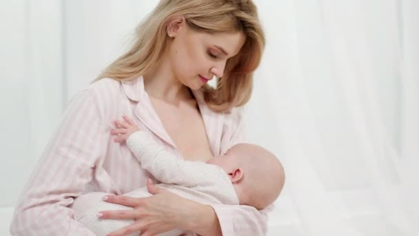 A young caucasian mother breastfeeds a newborn baby sitting on a bed in bedroom. — Stock Video