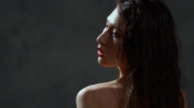 Uzun kıvırcık kıvırcık siyah saçlı, göğüs portresi, çıplak omuzlu güzel bir genç kadın. Güzel doğal ten. Çıplak makyaj. Kırmızı dudaklar ve zarif eller. Kız gri bir arkaplanda poz veriyor.