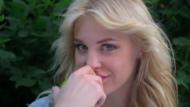 Portretul unei tinere femei frumoase cu păr blond ondulat împotriva unui parc. Față frumoasă, ochi albaștri și buze lucioase. Fata râde jucăuș și ține un măr verde în mâini . — Videoclip de stoc