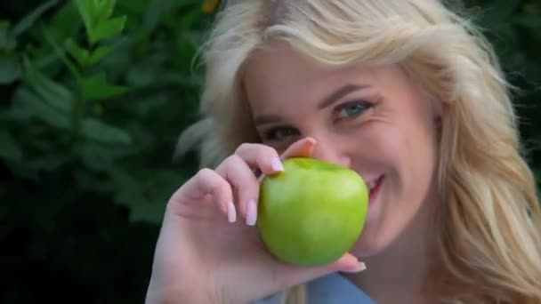 Retrato de una joven hermosa mujer con el pelo rubio rizado contra un parque. Hermosa cara, ojos azules y labios deliciosos. La chica ríe juguetonamente y sostiene una manzana verde en sus manos. — Vídeos de Stock