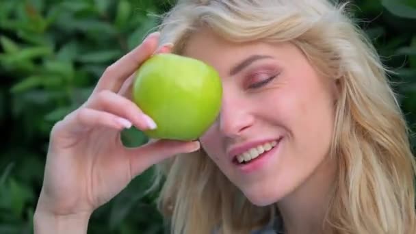 Portrét mladé krásné ženy s kudrnatými blond vlasy proti parku. Krásná tvář, modré oči a svůdné rty. Dívka se hravě zasměje a drží v rukou zelené jablko.
