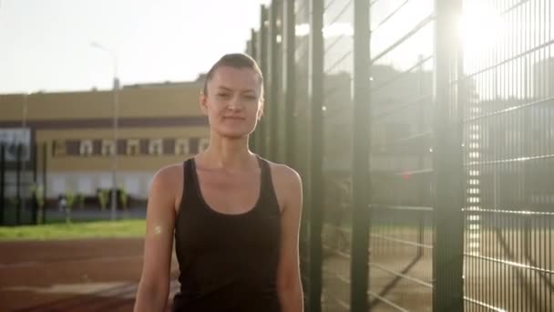 Eine junge schöne kaukasische Brünette posiert in Sportkleidung vor dem Hintergrund des Sportplatzes. Aktiver Lebensstil. — Stockvideo