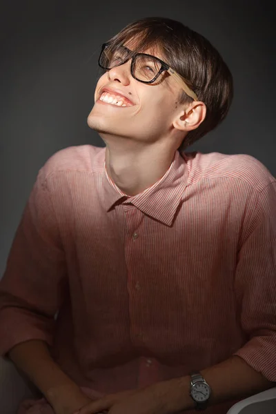 Ritratto di astuto divertente giovane caucasico che indossa occhiali, camicia in una striscia seduta al chiuso con viso felice e sorridente su sfondo grigio scuro . — Foto Stock